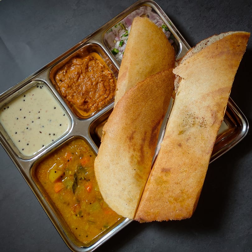 Two golden-brown dosas with three Indian chutneys and a vegetable curry in a metal tray.